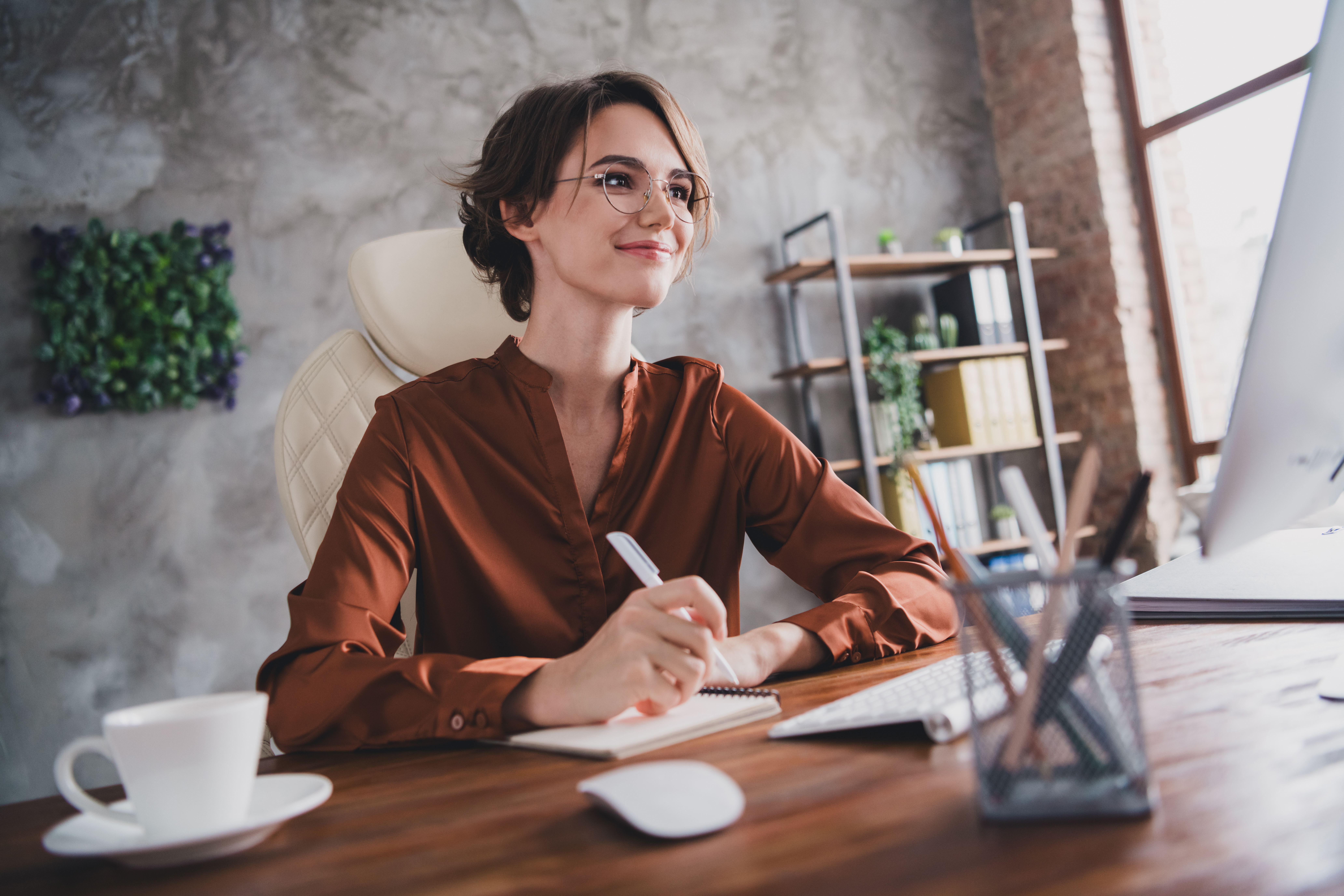 Osoba siedzi przy biurku w nowoczesnym biurze, trzyma długopis i robi notatki w notesie, patrząc w stronę monitora stojącego przed nią. 