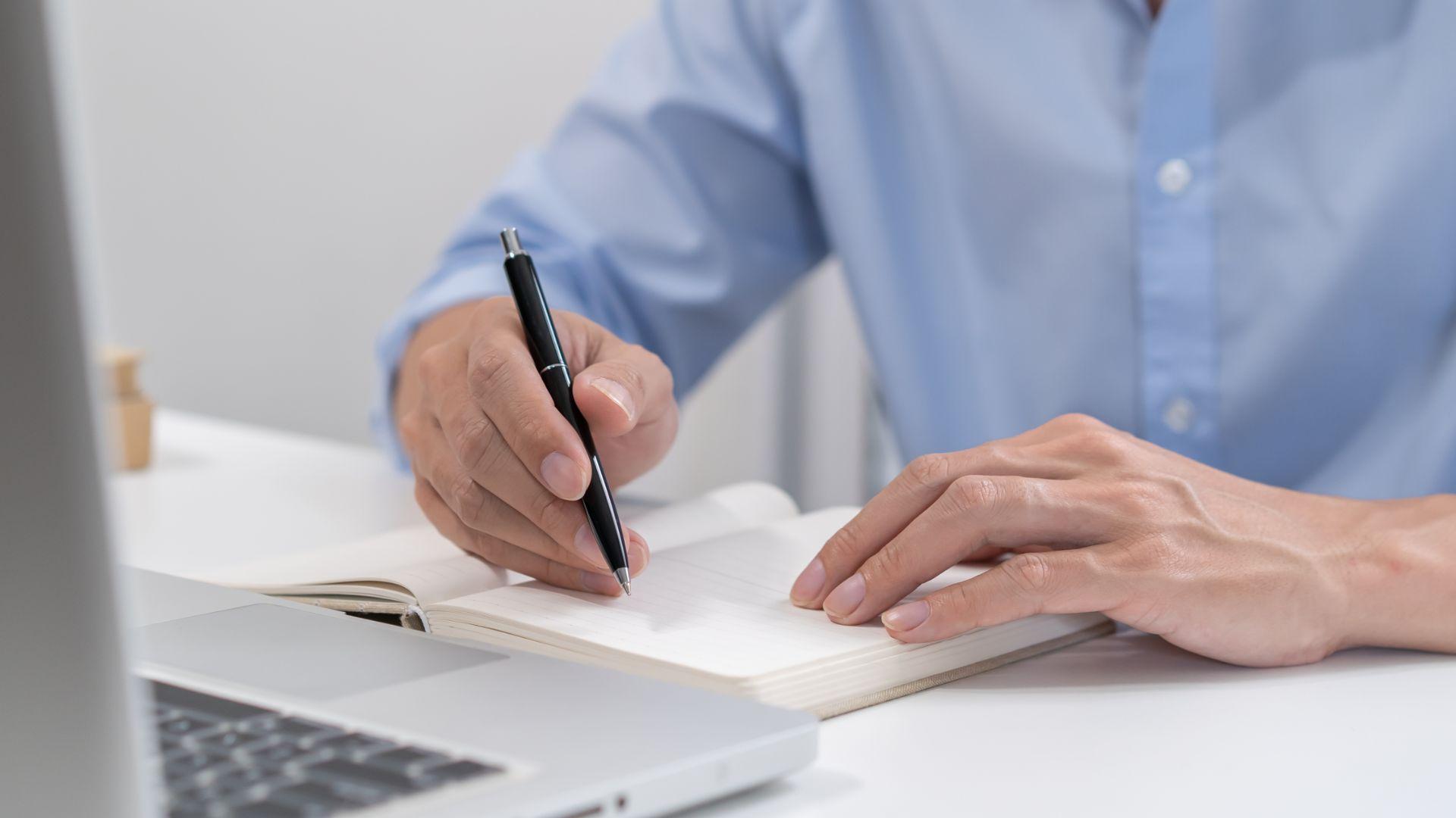 Osoba robi notatki w notesie przy laptopie