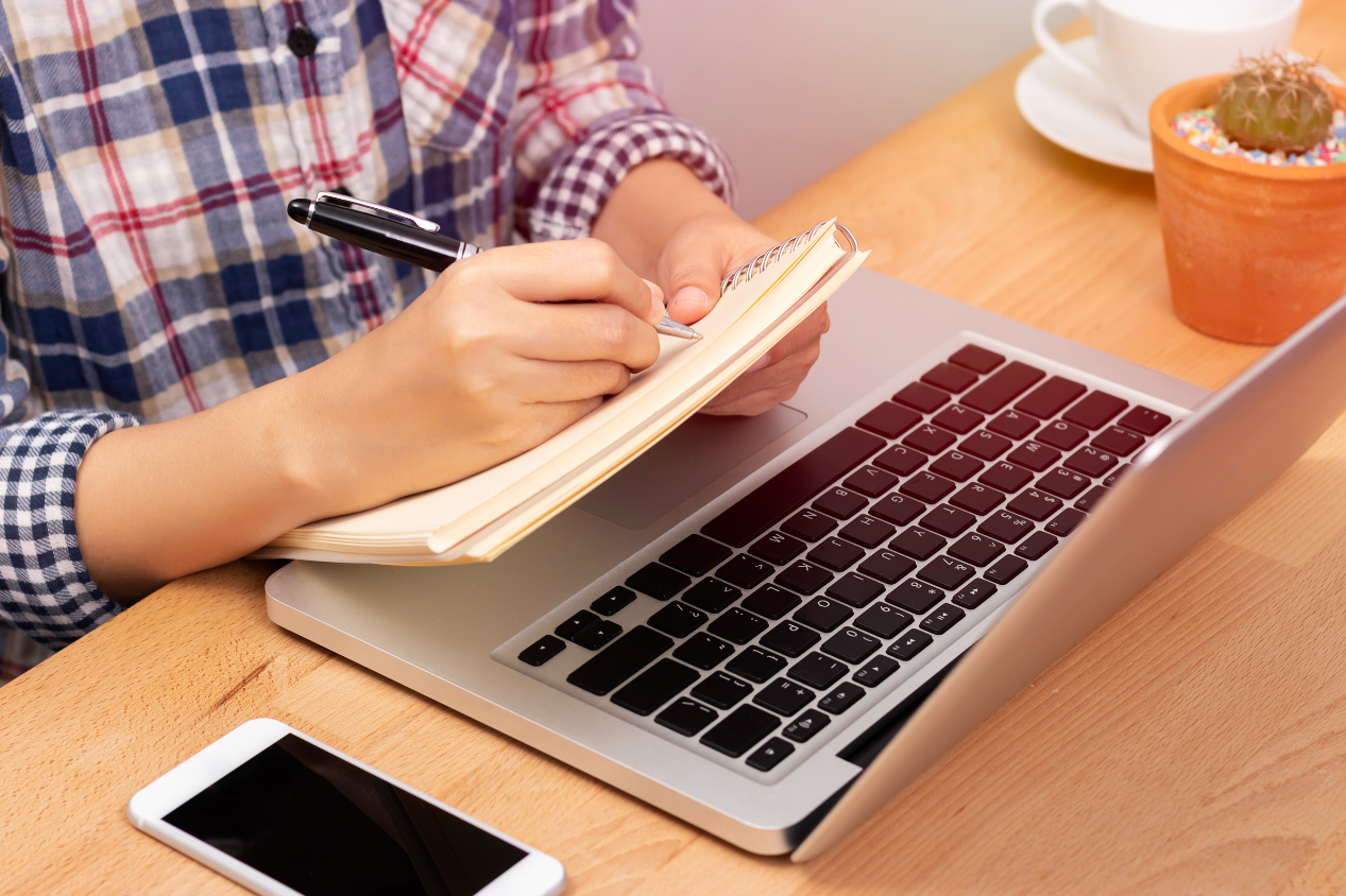 Osoba notuje w zeszycie spoglądając na ekran laptopa